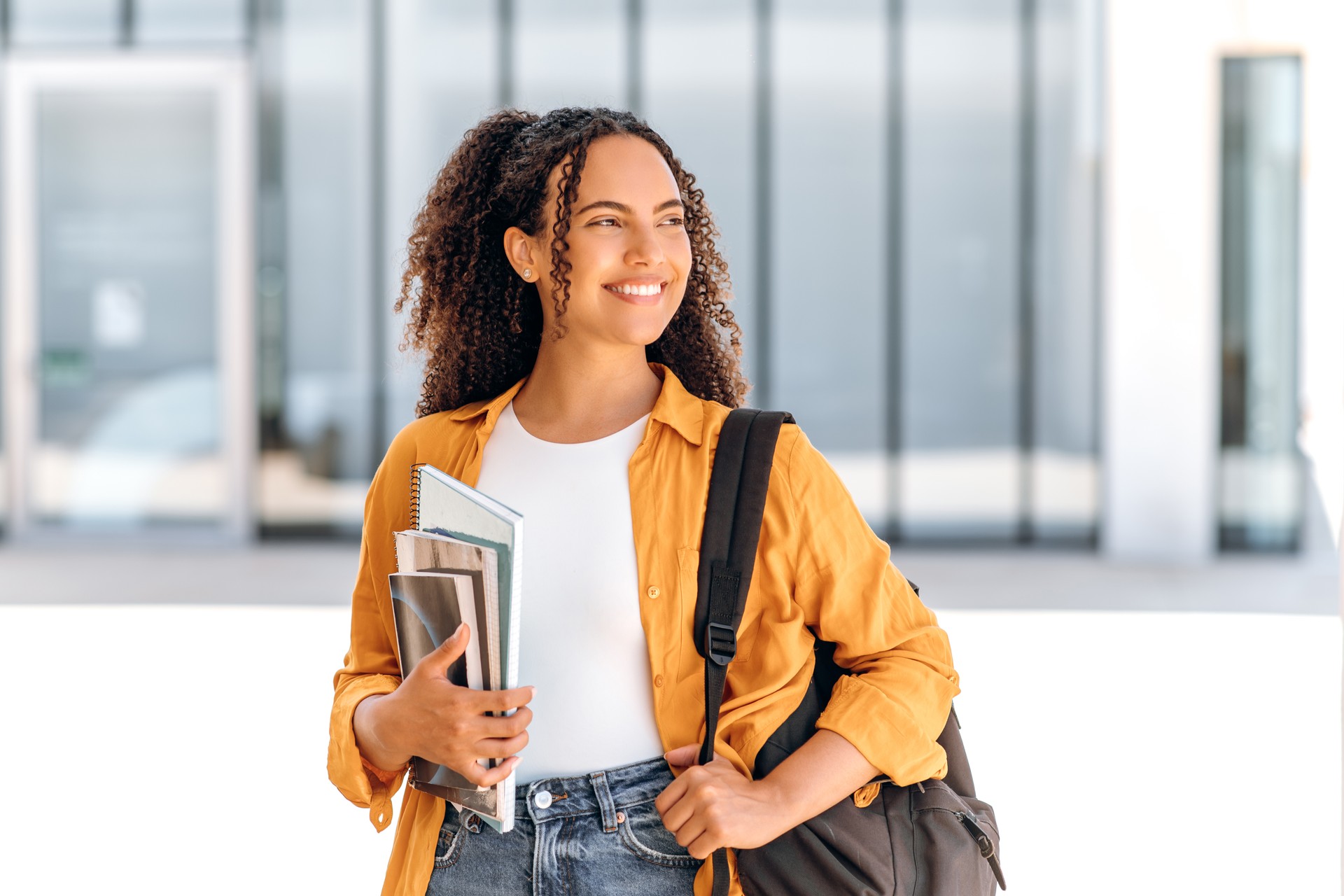 Estudante feliz. Estudante do sexo feminino, de nacionalidade brasileira ou hispânica, com mochila, segurando livros e cadernos na mão, fica perto do campus universitário, desvia o olhar e sorri